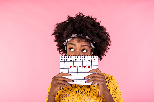 Portrait of a funny young African American girl in hiding behind a menstrual periods calendar and looking away at copy space isolated over pink background. Female Period calendar